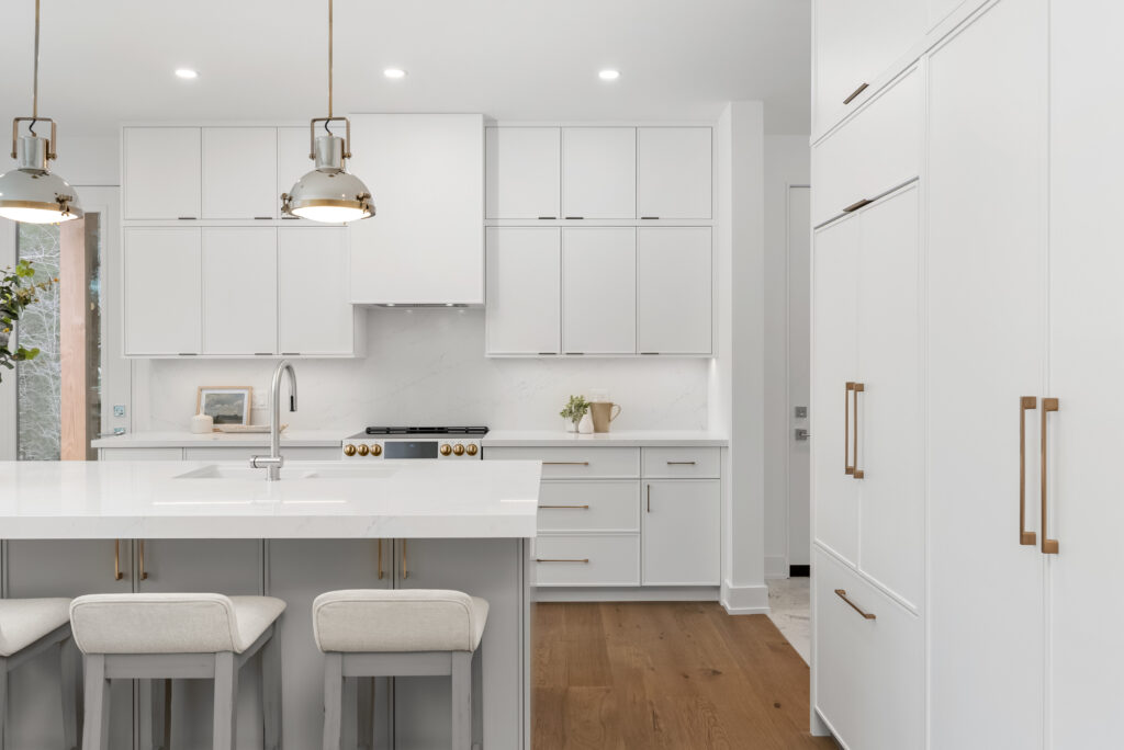 Modern white kitchen with gold hardware, pendant lights, an island with two cushioned stools, built-in appliances, and wood flooring. The space is bright with minimalist décor and sleek cabinetry.
