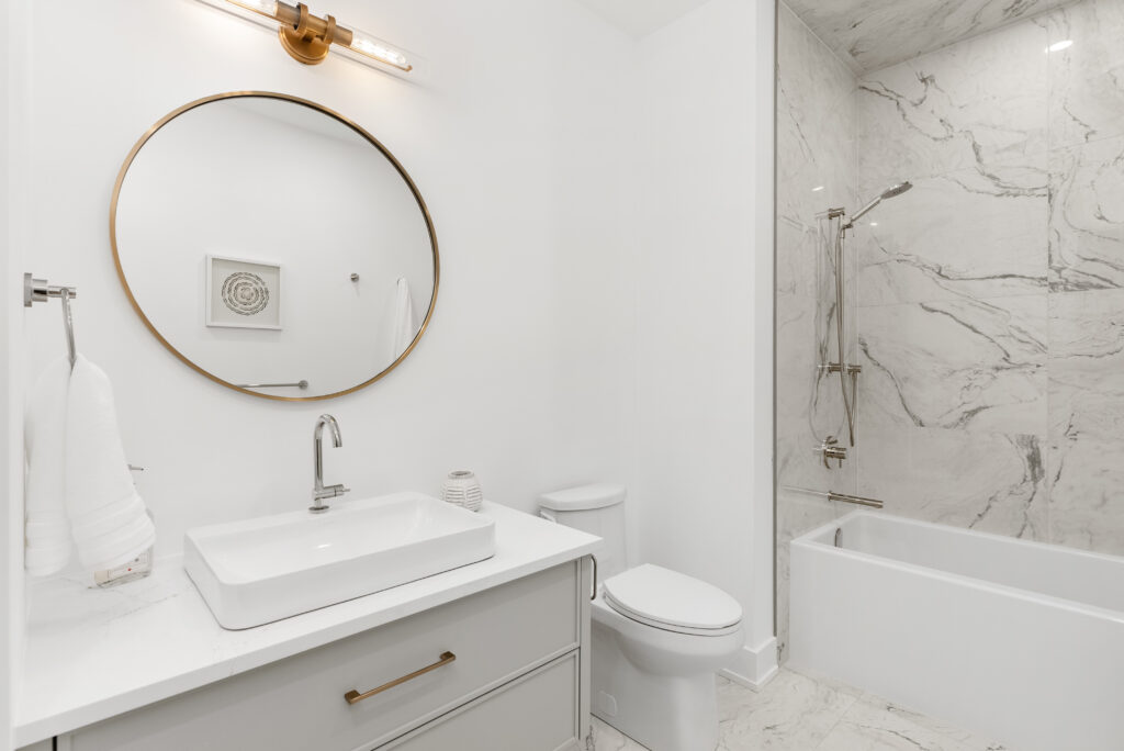 Modern bathroom with a round mirror above a white sink, a toilet, and a bathtub with a marble-tiled shower. The decor is minimal, featuring white walls and a gold-accented vanity and light fixture.