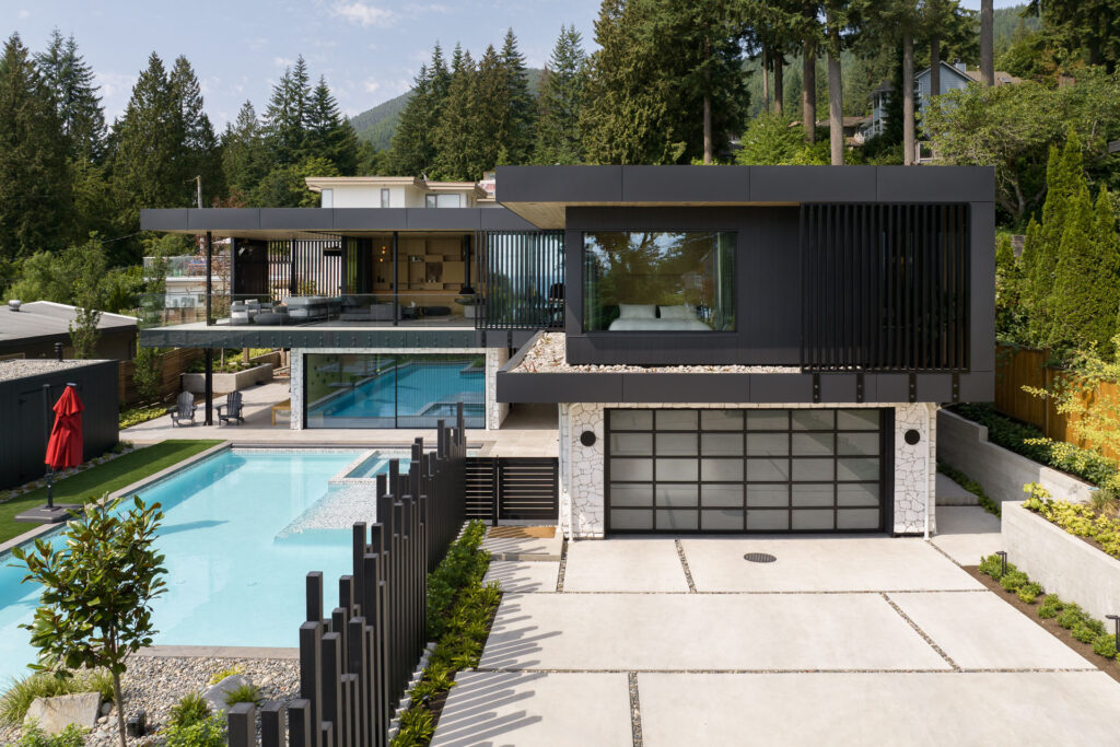 A modern two-story house with large glass windows, an outdoor swimming pool, and surrounding greenery. The home features a flat roof, a spacious patio, and a garage with a glass-paneled door. Trees and mountains are in the background.