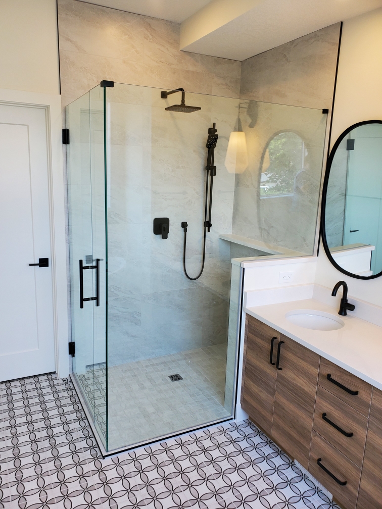 A modern bathroom with a glass-enclosed shower, black fixtures, a wood vanity with a white countertop and black faucet, an oval mirror, and patterned floor tiles.