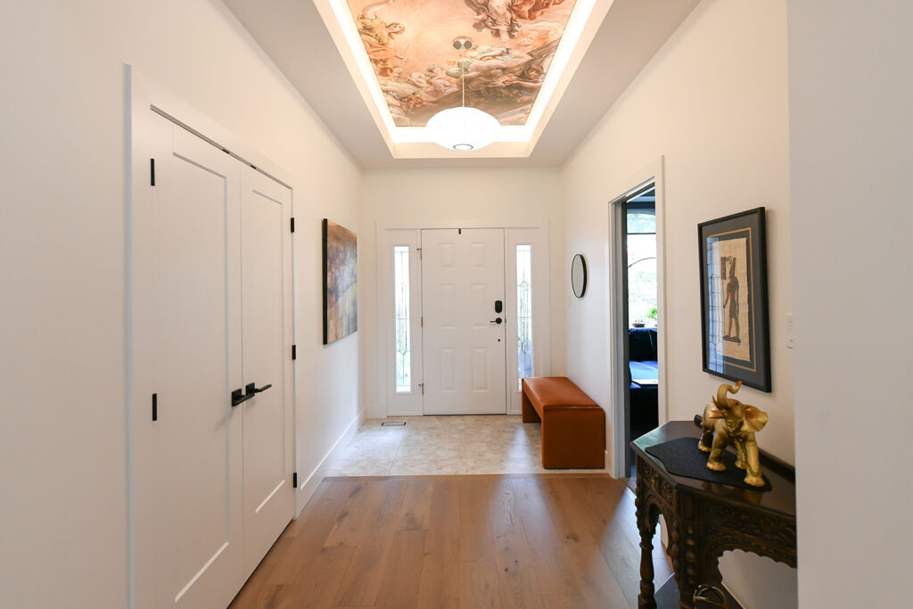 A bright hallway with white walls, a wooden floor, and a decorative ceiling mural. There’s a white front door, a brown bench, art on the walls, and a small table with a gold dog figurine on the right.