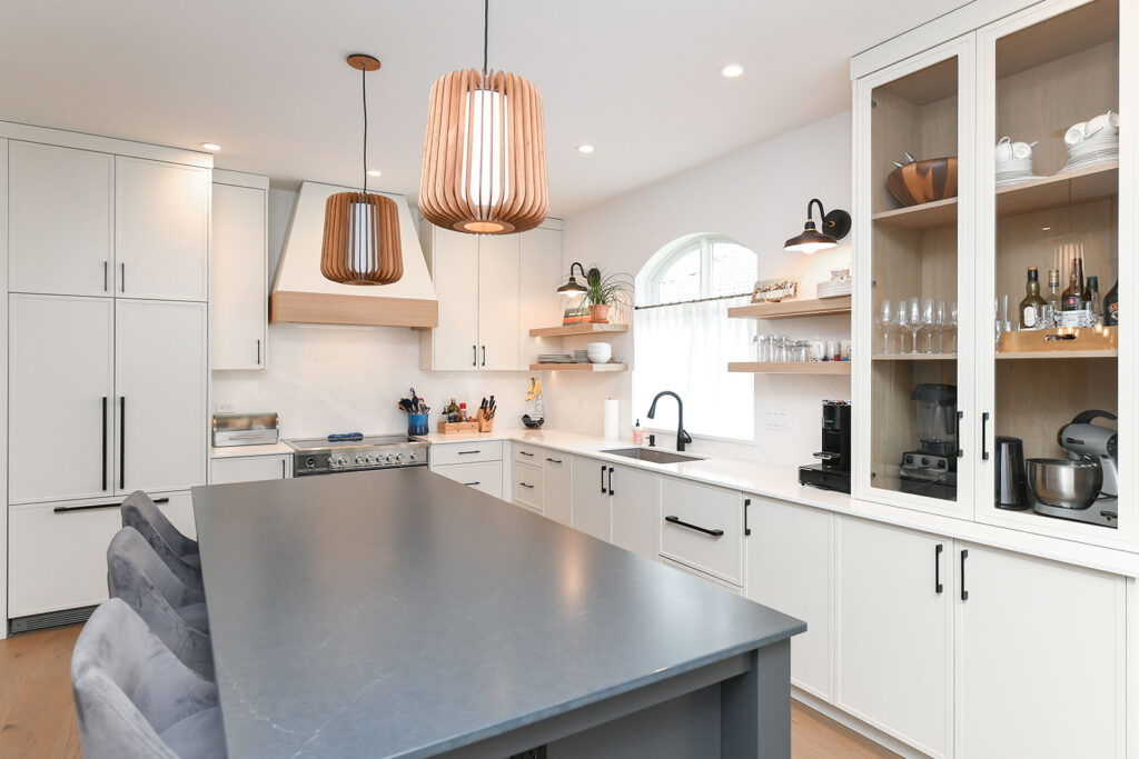 A modern kitchen with white cabinets, a gray island with bar stools, pendant lights, open shelves, a stove, sink under a window, and glass-front cabinets displaying glassware and kitchen appliances.