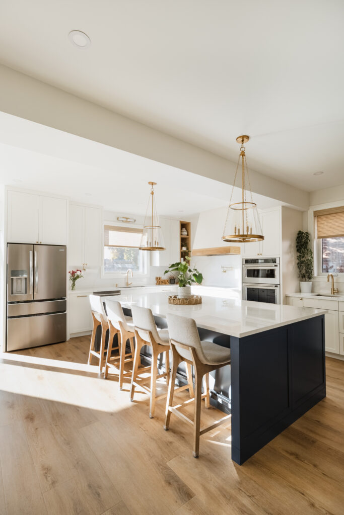 Bright, modern kitchen with a large navy-blue island, white countertops, four wooden and upholstered chairs, stainless steel appliances, pendant lights, light wood flooring, and white cabinetry.