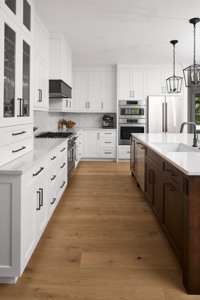 Modern kitchen with white cabinets, stainless steel appliances, a large island with a dark wood base and white countertop, pendant lights, glass cabinet doors, and light wood flooring.