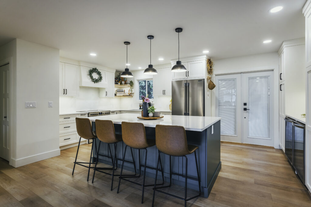 Modern kitchen with a large island, four brown bar stools, pendant lights, white cabinets, stainless steel appliances, and wood flooring. Decor includes a plant, flowers, and a wreath on the wall.