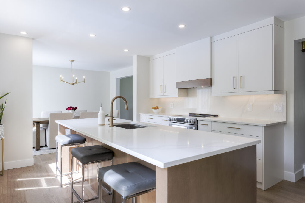 Modern kitchen with white cabinets, a large island with a built-in sink, gray cushioned stools, stainless steel appliances, and a view of a dining area with a light fixture in the background.