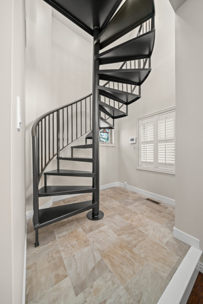 A black metal spiral staircase stands on a tiled floor in a bright room with light-colored walls and a window with white shutters.