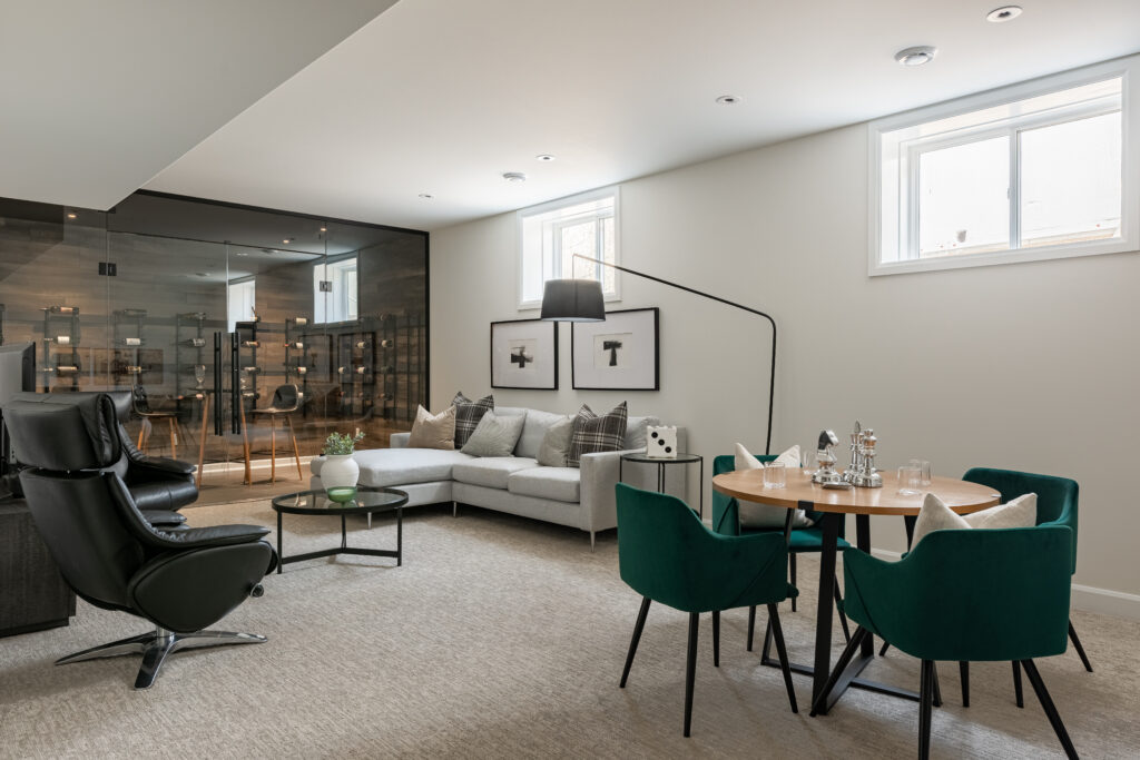 Modern living room with a gray sectional sofa, black armchair, round coffee table, and a dining area with a wooden table and four green velvet chairs. Large windows and glass-walled room add brightness to the space.