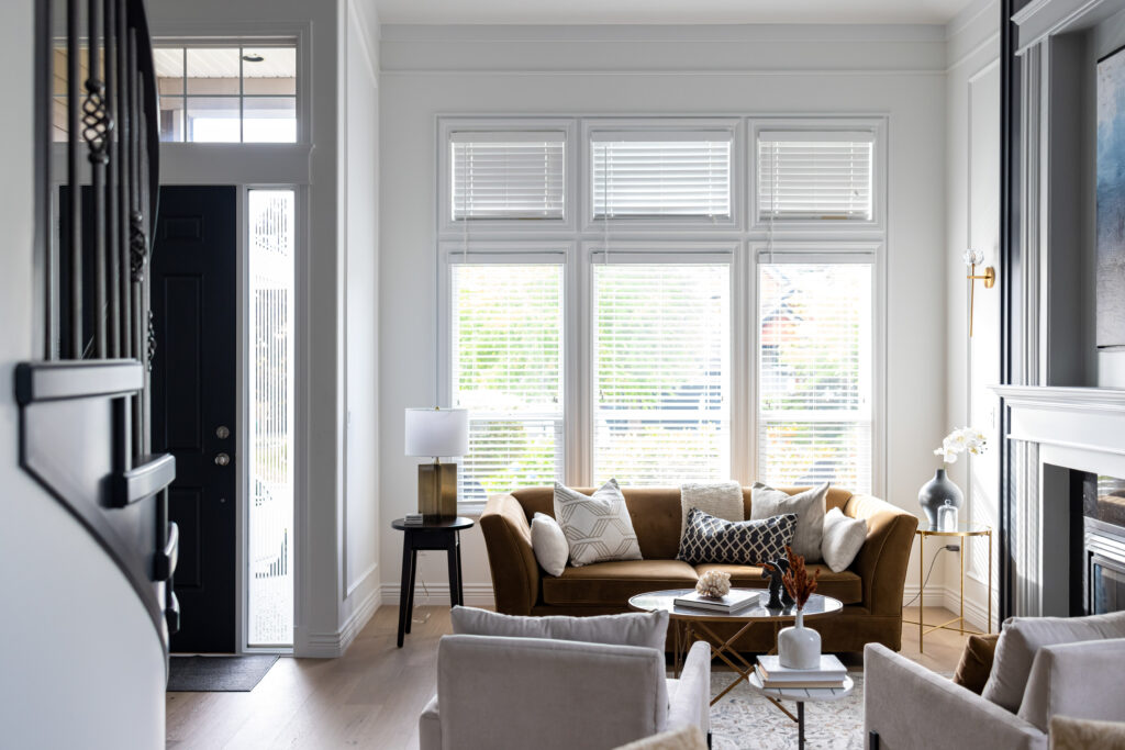Bright living room with a brown sofa, two beige chairs, coffee table, and large windows with white blinds letting in natural light. Modern decor includes pillows, lamps, and a small plant.