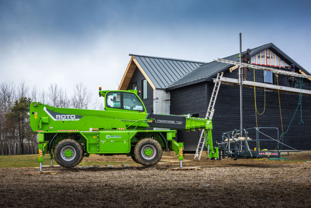 A bright green ROTO telehandler is parked beside a dark wooden house under construction, with a worker on scaffolding installing siding near the roof and tools and ladders nearby on a cloudy day.