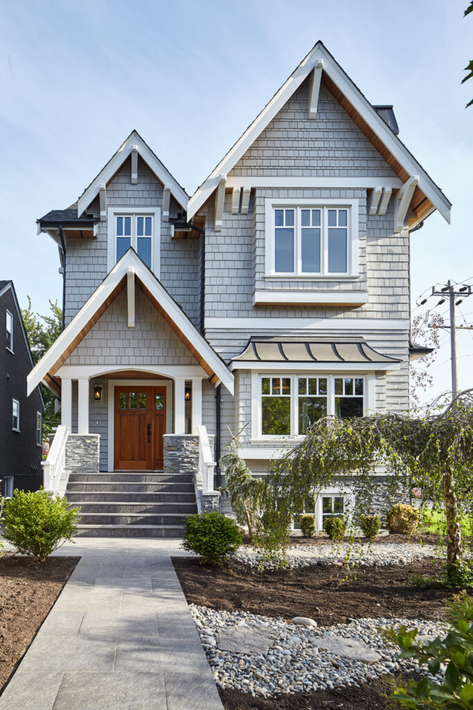 A two-story gray house with white trim, a peaked roof, and a wooden front door. The entrance has stone steps and railings, and the landscaped yard features shrubs and a stone walkway.