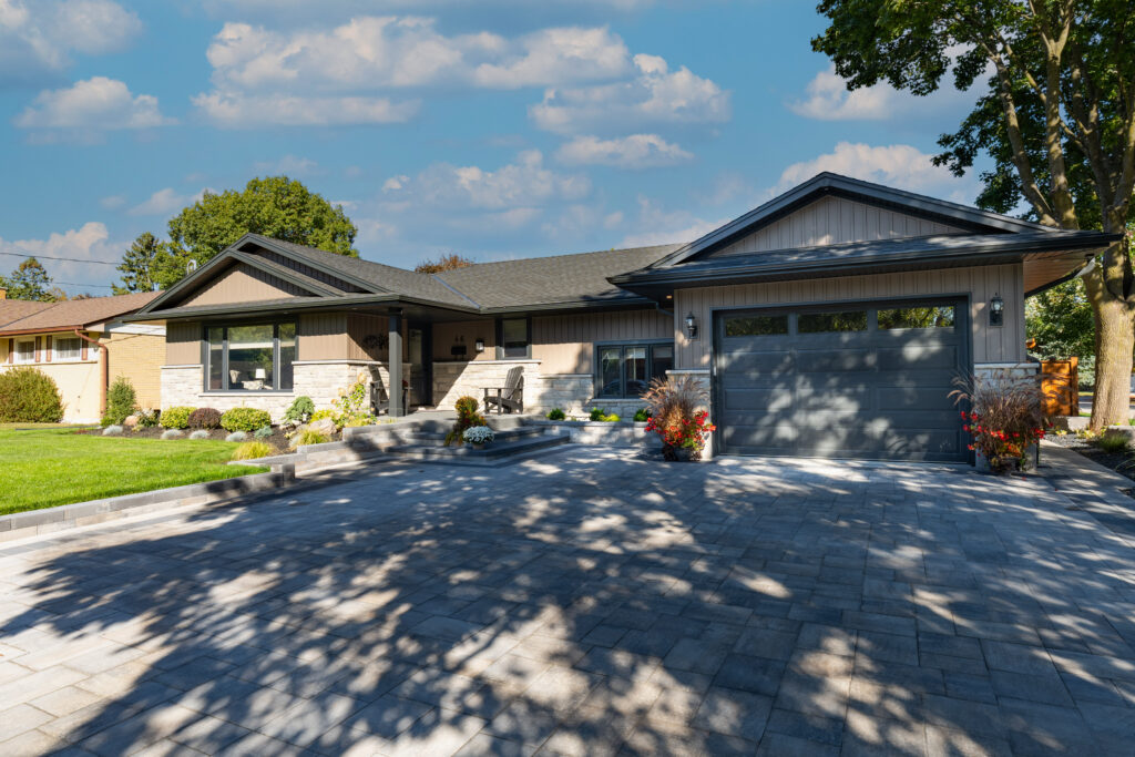 A modern single-story house with a gray garage, stone walkway, landscaped front yard, and potted plants, under a blue sky with scattered clouds. Shadows from nearby trees fall across the driveway.