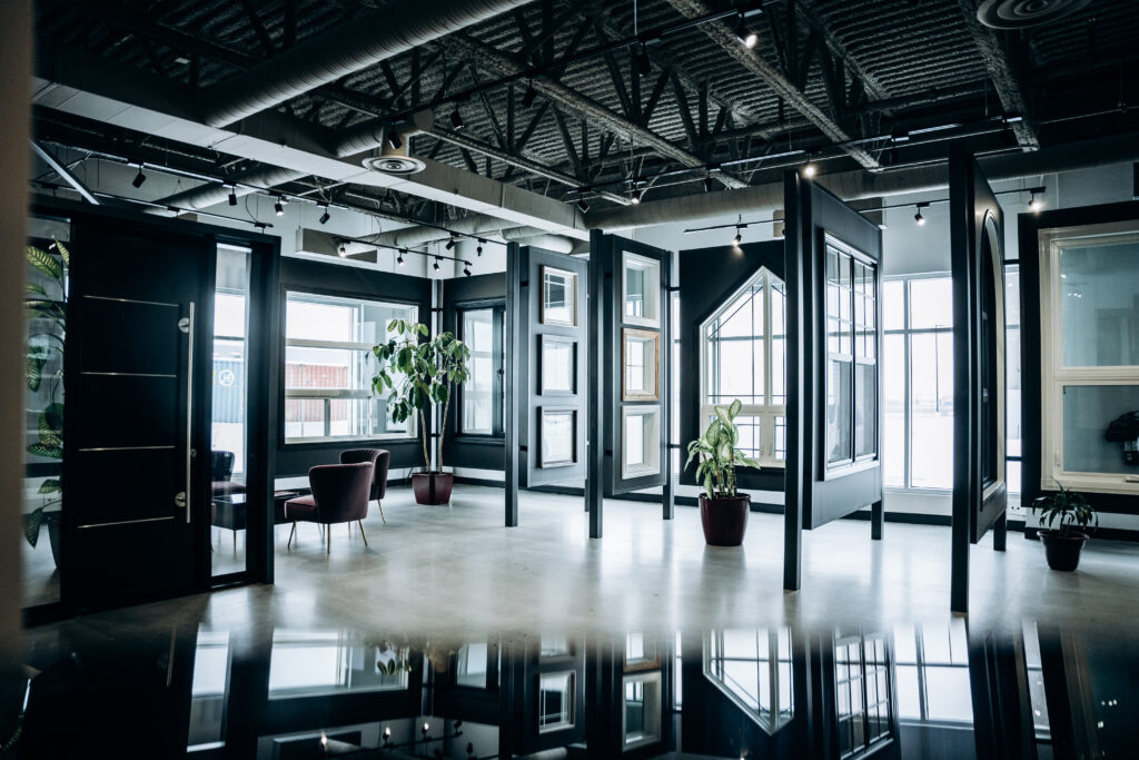A modern, open office space with large windows, potted plants, and several door frames standing upright. Natural light fills the area, which features exposed ceilings and minimalistic furniture, including two lounge chairs.