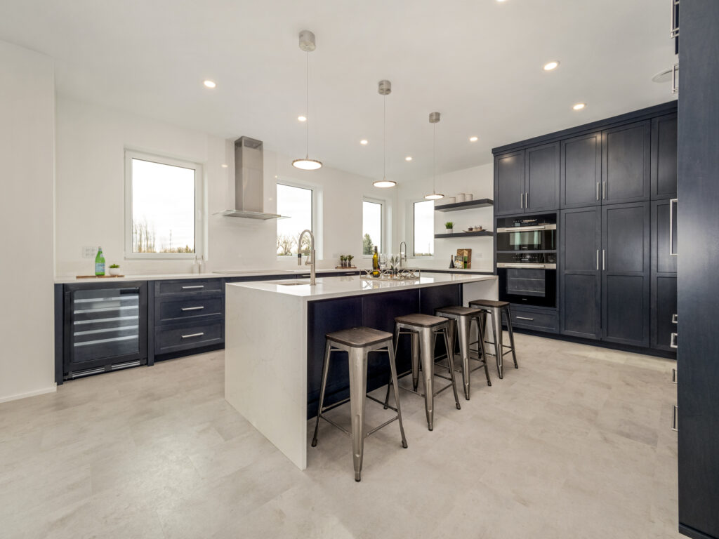 Modern kitchen with navy blue cabinets, a large white island with four metal stools, built-in appliances, pendant lights, and large windows letting in natural light. The space is clean and spacious with a minimalist design.