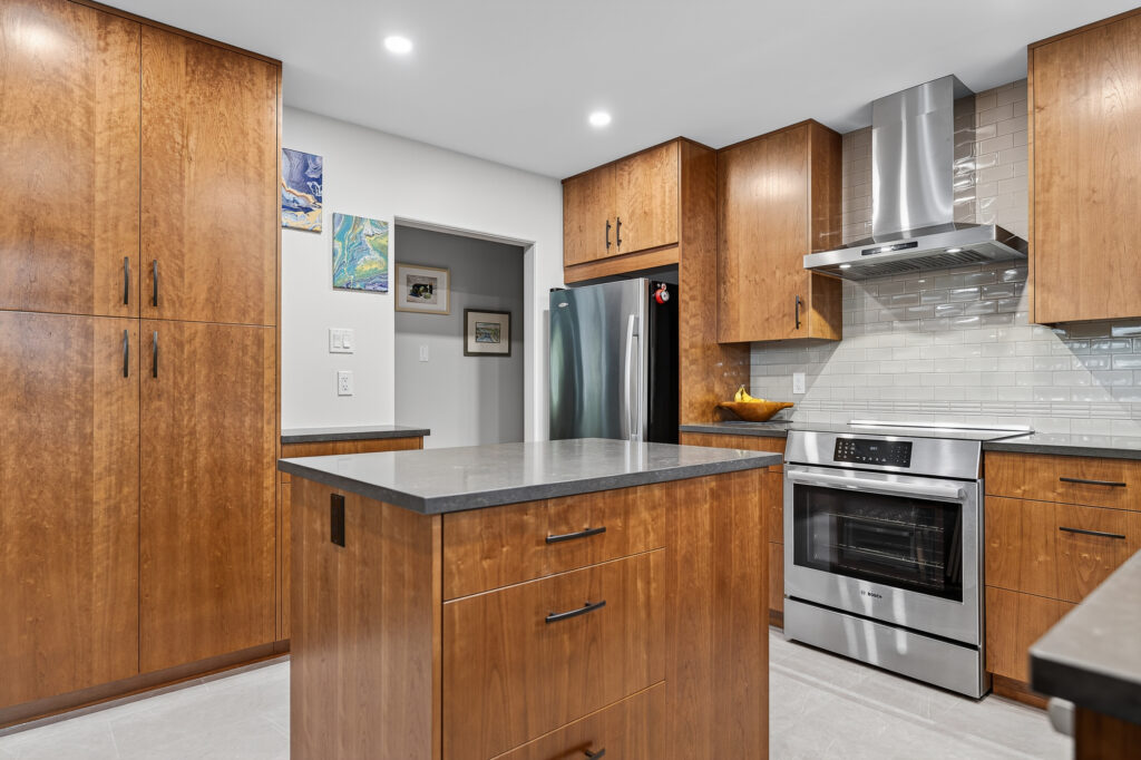 Modern kitchen with wooden cabinets, a central island with drawers, stainless steel appliances including a fridge and oven, a range hood, light tile flooring, and light-colored backsplash tiles. Bright lighting and wall art are visible.
