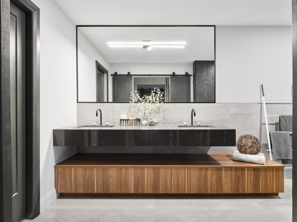 Modern bathroom with a large rectangular mirror, double sink with black countertop, wooden cabinet below, decorative accents, and a towel ladder on gray tiled floor and walls. Neutral tones and minimalist design.