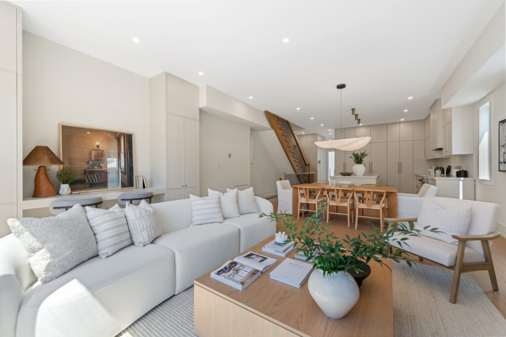 A modern, bright open-concept living area with a white sectional sofa, wooden coffee table, neutral decor, dining area, and kitchen. Stairs lead upstairs, and natural light fills the space through large windows.