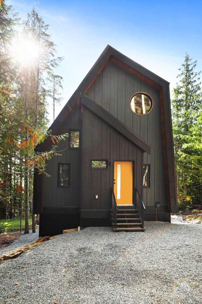 A modern, dark gray, A-frame house with a bright yellow door and a round window, set among tall trees under a sunny blue sky, with gravel covering the front yard.
