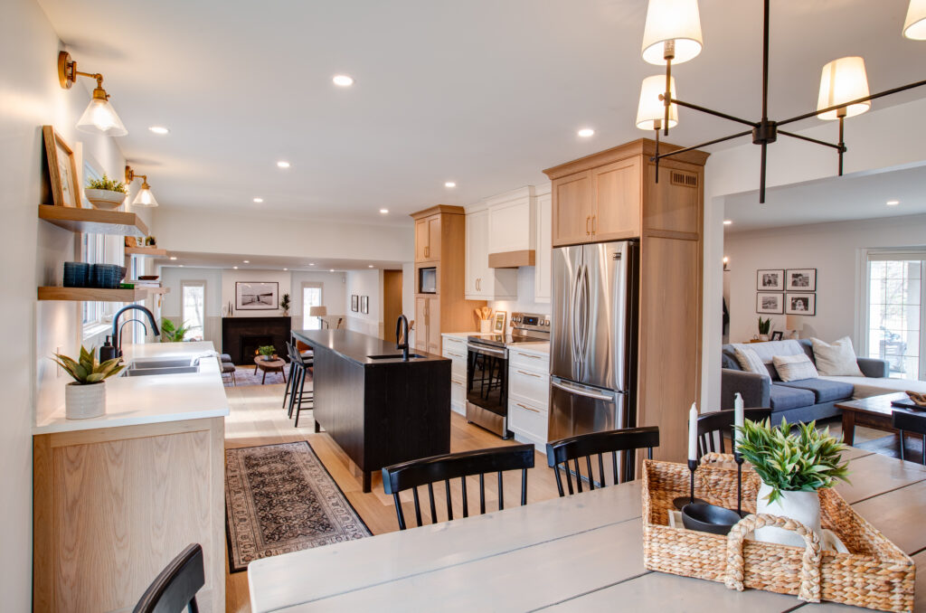 Open-concept kitchen and dining area with light wood cabinets, stainless steel appliances, a kitchen island, shelves with plants, a dining table with black chairs, and a living room visible in the background.