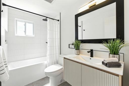 Modern bathroom with a white bathtub and shower, toilet, and a vanity with a sink. A large mirror hangs above the sink, and two potted plants decorate the countertop. Light enters through a small window.