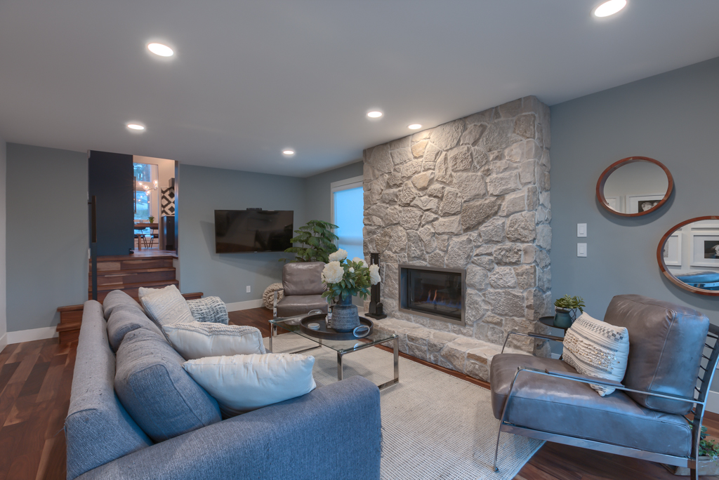A modern living room with gray sofas, an armchair, a glass coffee table, and a stone fireplace. The space features hardwood floors, recessed lighting, and neutral decor with green plants and wall art.