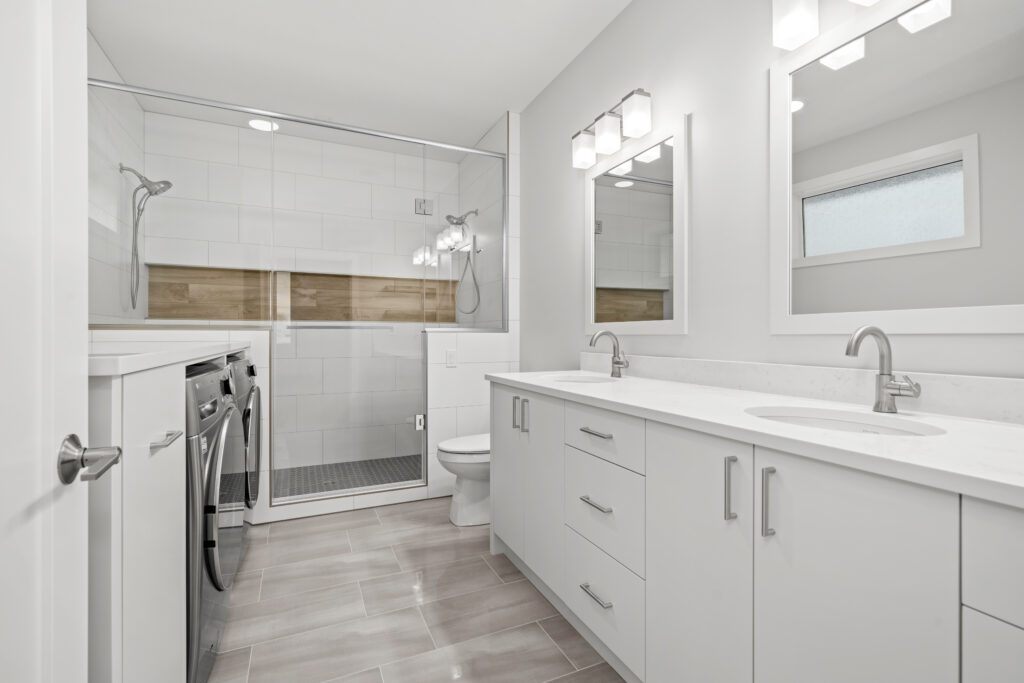 Modern bathroom with white cabinets, a double-sink vanity, large mirrors, a glass-enclosed shower, washing machine, dryer, and a frosted window. The space is bright with sleek, minimalist design.
