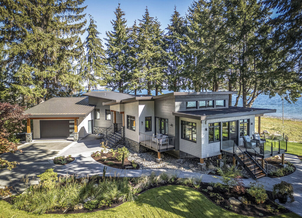 Modern two-story house with large windows, a porch, and an attached garage, surrounded by tall trees and landscaped gardens, with a view of water and blue sky in the background.