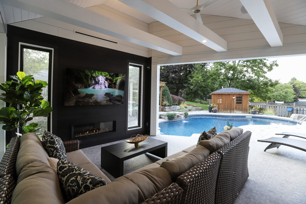 A covered patio with a wicker sectional sofa, coffee table, TV above a fireplace, and a view of a backyard pool, garden shed, and trees through large windows and open walls.