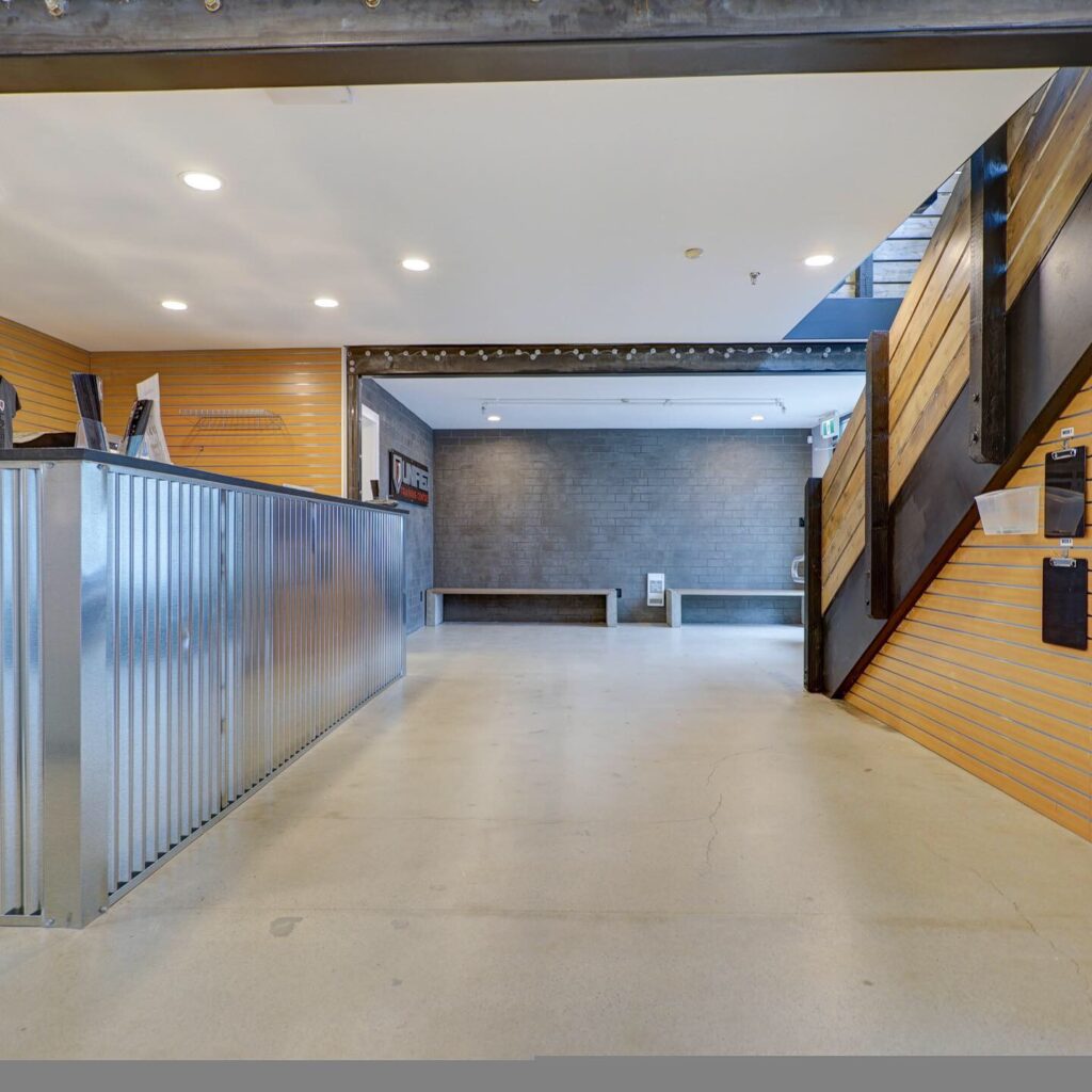 A modern, industrial-style interior with a corrugated metal reception desk on the left, wood-paneled walls, a staircase on the right, and concrete floors extending to a gray brick wall at the back.