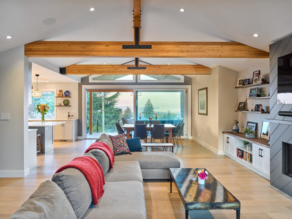 Modern open living space with a gray sectional sofa, red throw, rustic wood beams, dining area with large windows, built-in shelves, and a view of trees and the outdoors. Bright, airy, and stylish interior design.