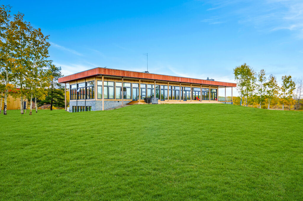 A modern, single-story building with large glass windows sits atop a green, grassy hill under a bright blue sky, surrounded by trees with autumn foliage.