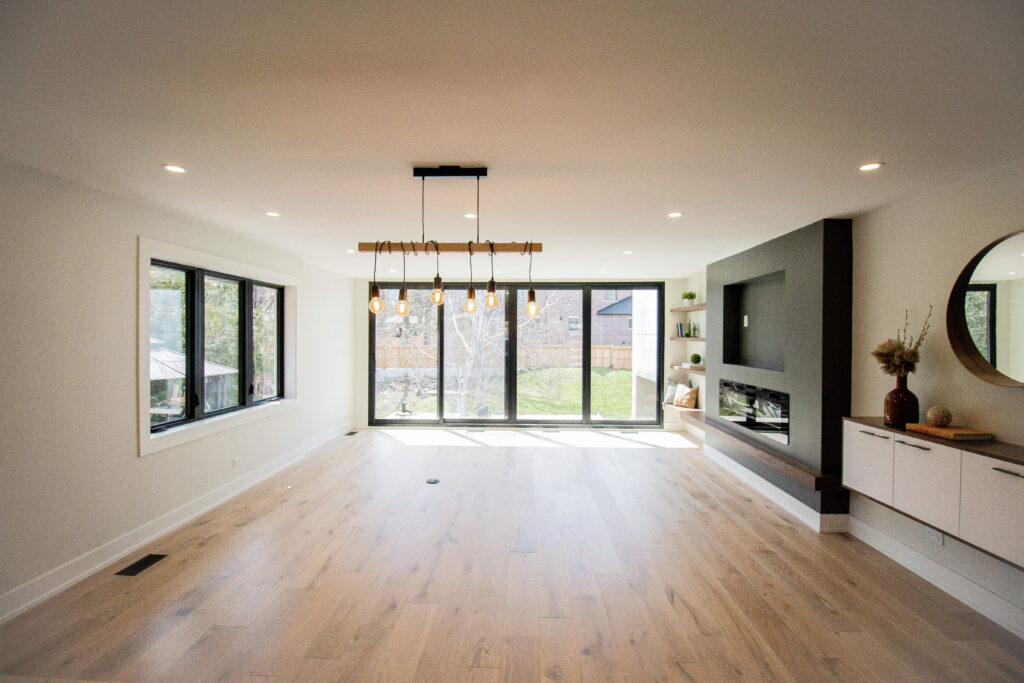 A modern, empty living room with large windows, light wood floors, a black accent wall with a built-in fireplace, and a contemporary light fixture hanging from the ceiling.