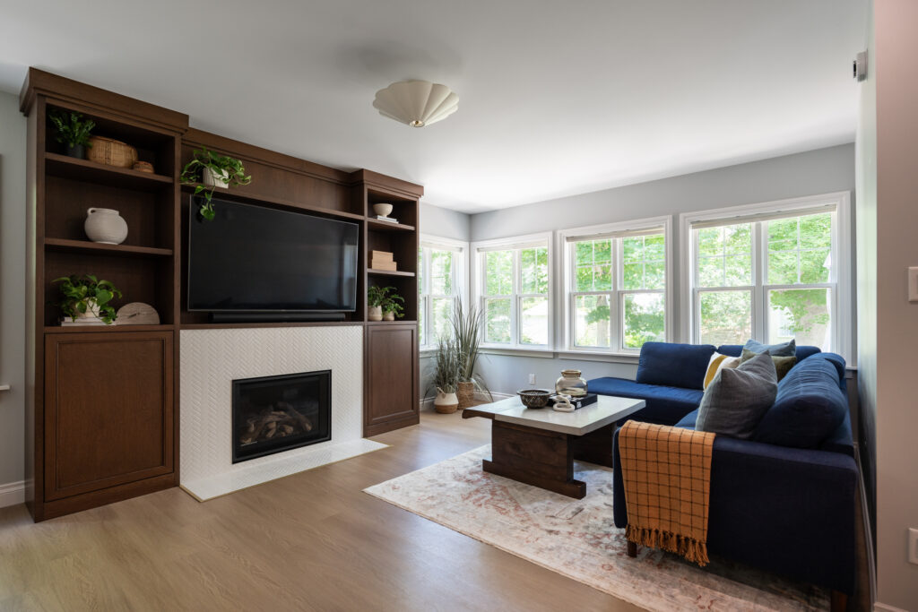 A cozy living room with a navy sofa, wooden coffee table, and built-in wooden shelves around a fireplace and large TV. Large windows let in natural light, and green plants decorate the space.