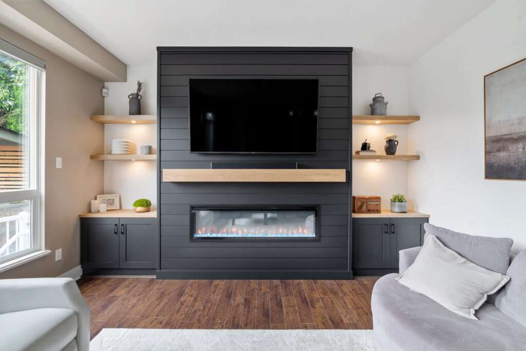 Modern living room with a black shiplap fireplace wall, mounted TV, built-in electric fireplace, floating wooden shelves with decor, cabinets below, and cozy seating on hardwood flooring.