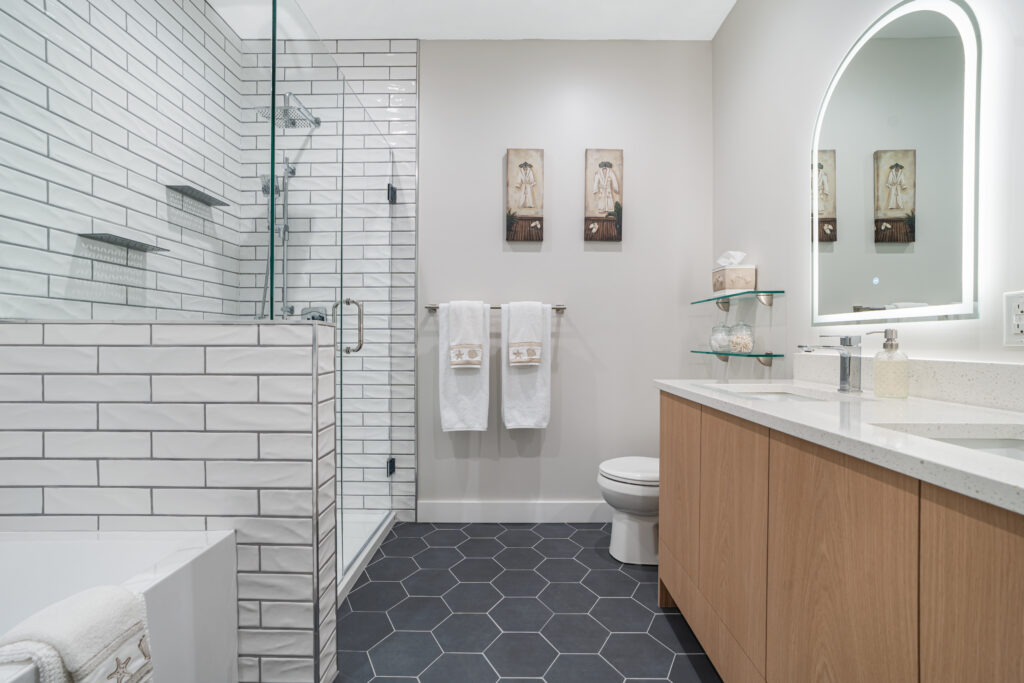 Modern bathroom with hexagonal dark floor tiles, a glass shower with subway tiles, a wooden vanity with white countertop, oval backlit mirror, toilet, towel rack, glass shelves with decor, and two wall art pieces.