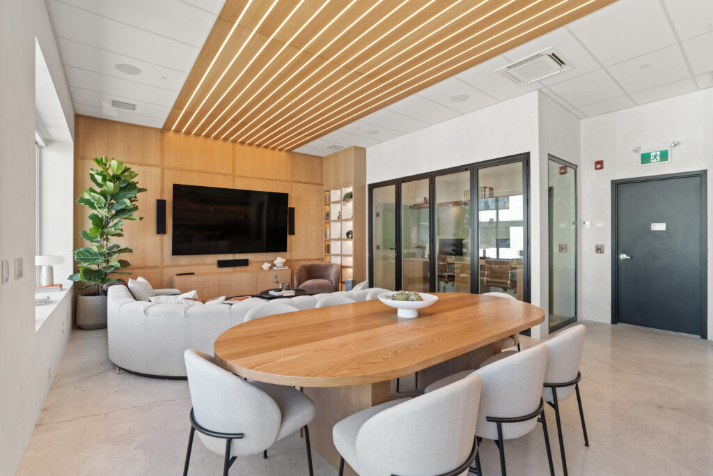 Modern office lounge with a wooden oval table, white cushioned chairs, a curved light gray sofa, large TV on a wooden wall, indoor plant, and glass-panel meeting rooms in the background. Warm lighting from wood-paneled ceiling.