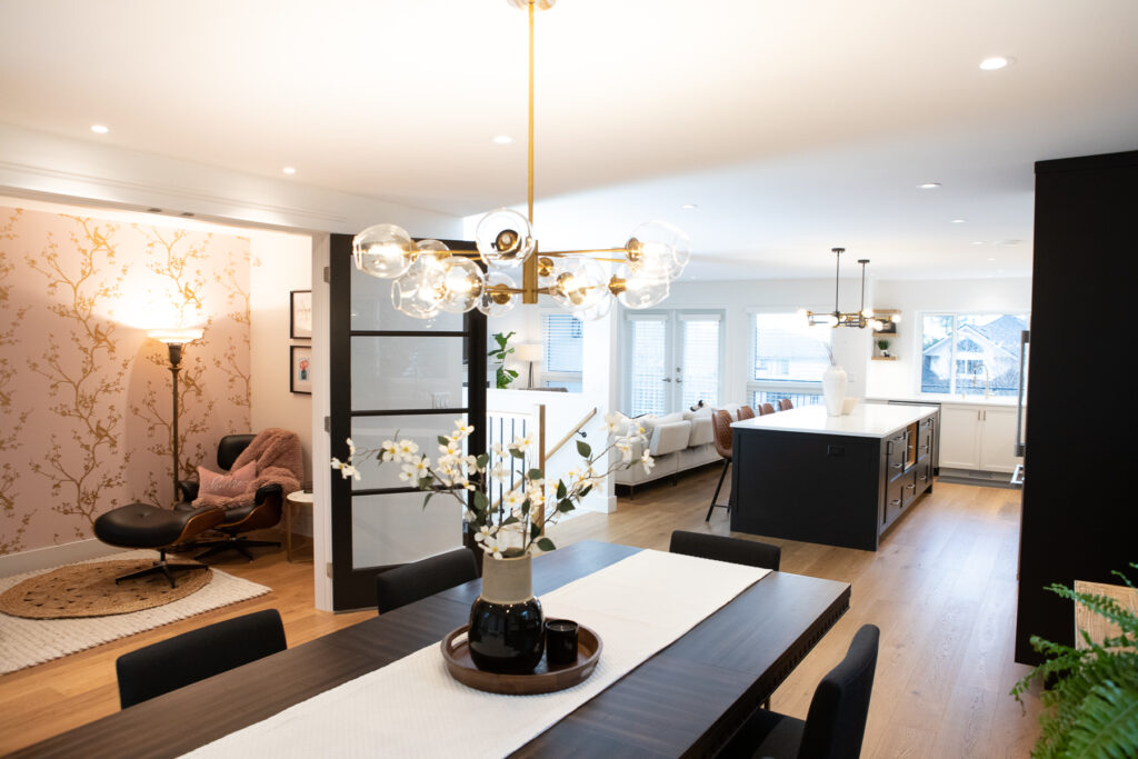 Modern open-concept living space featuring a dining table with a floral centerpiece, stylish light fixtures, a cozy lounge area with a chair and reading lamp, and a bright kitchen with large island and windows.