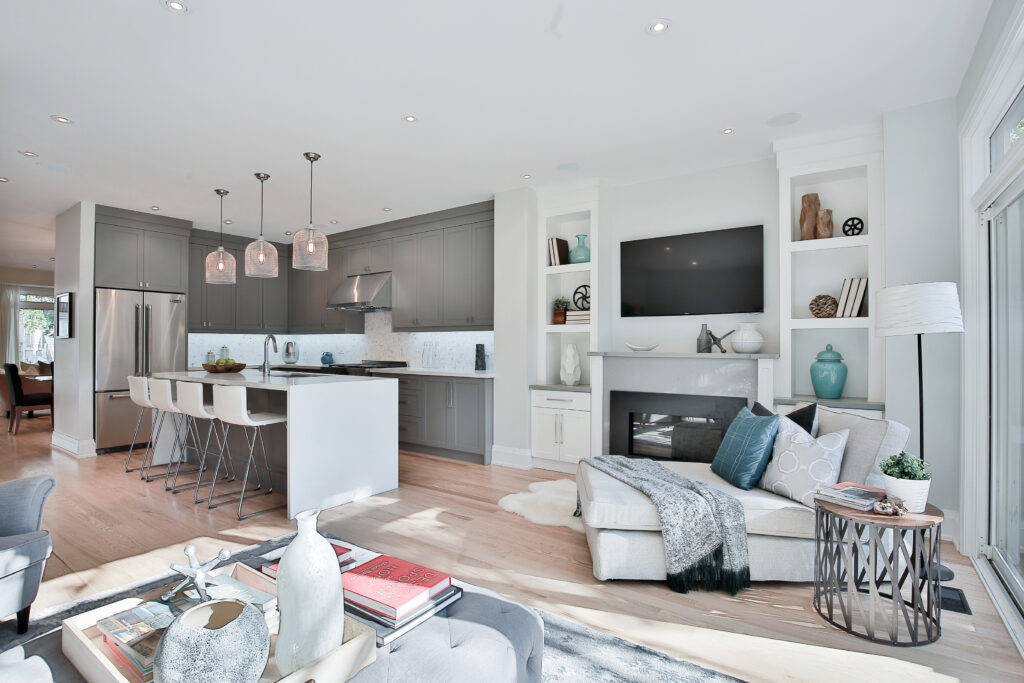 Modern open-concept kitchen and living area with light wood floors, gray cabinets, island with white barstools, built-in shelves, a fireplace, a wall-mounted TV, and cozy seating with decorative accents.