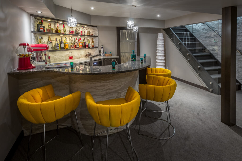 A modern home bar with a curved granite countertop, three bright yellow bar stools, shelves stocked with bottles and glasses, pendant lights above, and a staircase with glass railing in the background.
