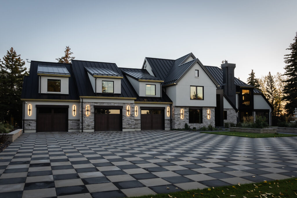 Modern two-story house with black metal roof, large windows, three garage doors, and stone accents. The spacious driveway is paved with a black and white checkerboard pattern. Tall trees surround the property at dusk.