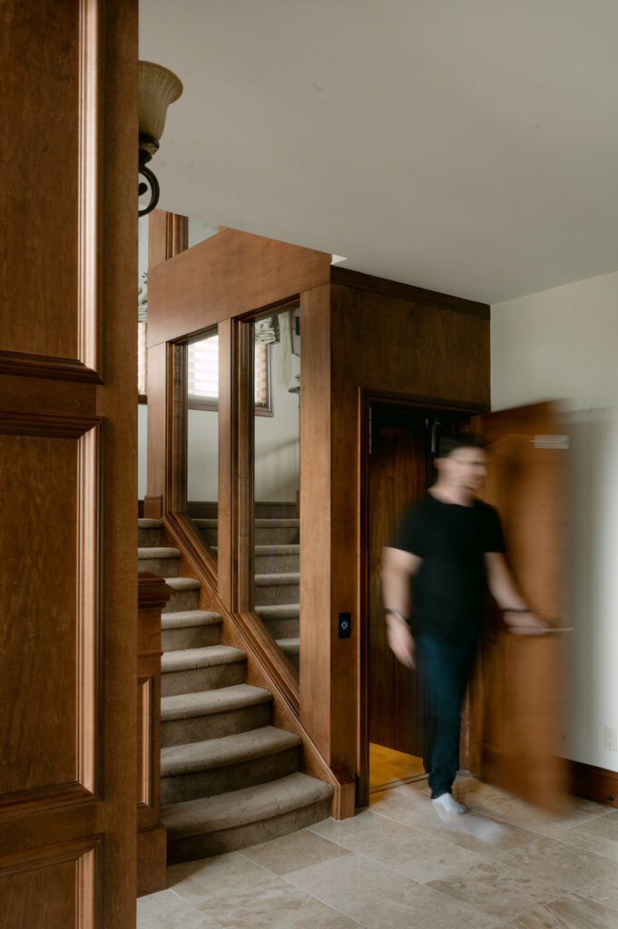 A man in a black shirt is blurred in motion as he opens a wooden door beside a staircase with brown wooden paneling and beige carpeted steps in a well-lit interior space.