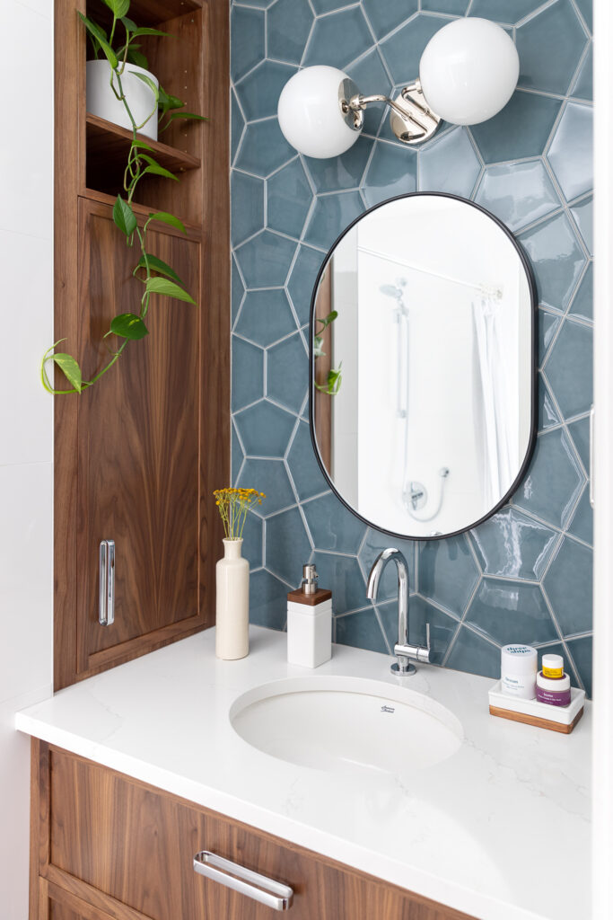 A modern bathroom vanity with a round mirror, blue hexagonal tile backsplash, white countertop, wood cabinets, plants, and minimal toiletries neatly arranged around the sink.