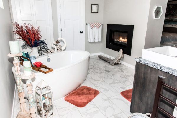 A bright bathroom with a white freestanding tub, marble floor tiles, coral-colored rugs, a black wall-mounted fireplace, decorative items, and candles. Towels hang by the door and a vanity sits to the right.