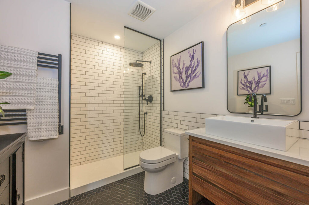 Modern bathroom with a glass-enclosed shower, white subway tile, black fixtures, a wooden vanity with a rectangular sink, large mirror, and framed purple botanical artwork on white walls.