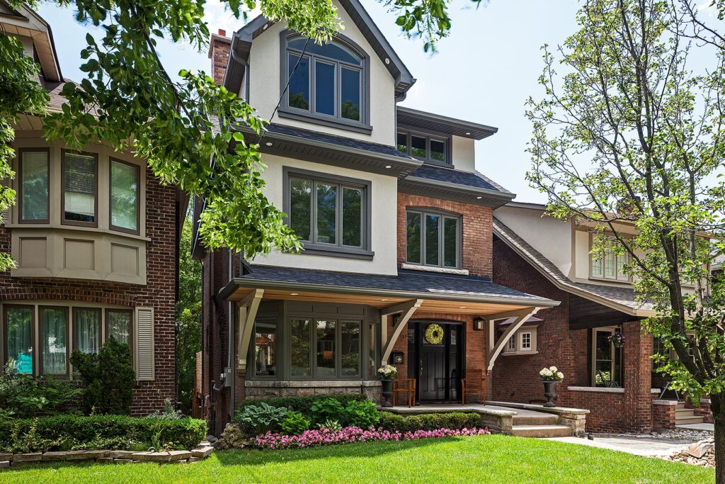 A modern three-story house with large windows, a covered porch, and a tidy front yard with green grass, shrubs, and flowers, situated between two similar brick homes on a sunny day.