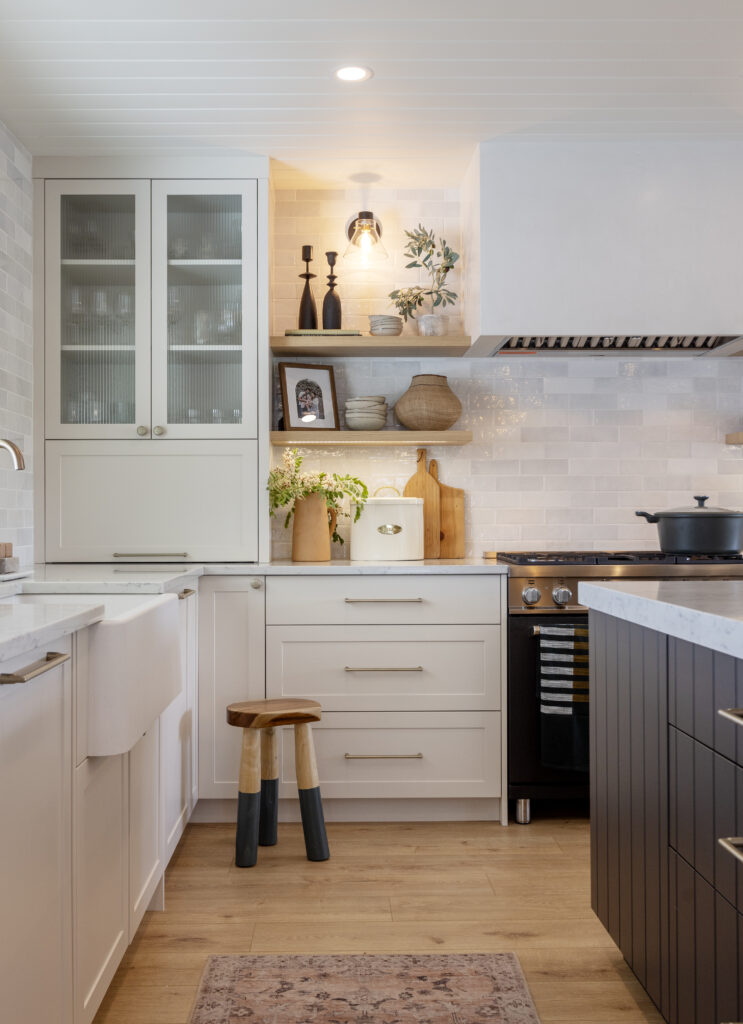 A modern kitchen with white cabinets, glass-fronted cupboards, wooden accents, a black stove, open shelves with decor, plants, a cutting board, and a small wooden stool on a light wood floor with a patterned rug.