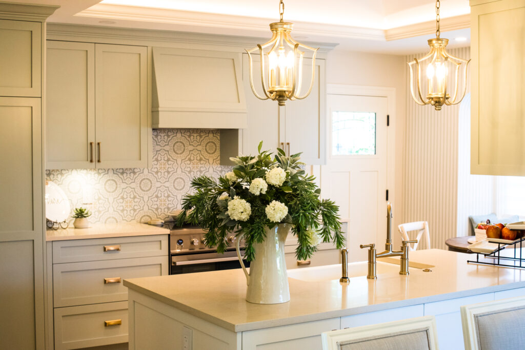 Bright, modern kitchen with light cabinets, patterned backsplash, gold fixtures, and pendant lights. A white pitcher with white flowers and greenery sits on the island countertop. A bowl of fruit is visible near the window.