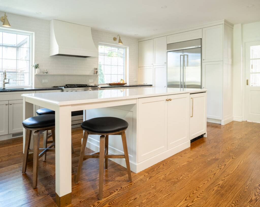 Bright, modern kitchen with white cabinets, a large island with two black stools, stainless steel refrigerator, built-in oven, range hood, and wooden floors. Sunlight streams in through large windows.