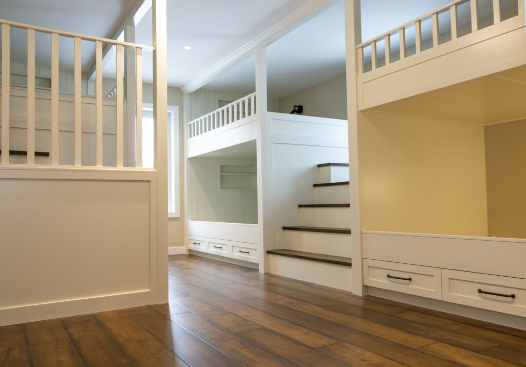 Interior view of a modern, spacious room with built-in bunk beds, wooden floors, white railings, drawers under beds, and a staircase leading to an upper sleeping area. Natural light enters through large windows.