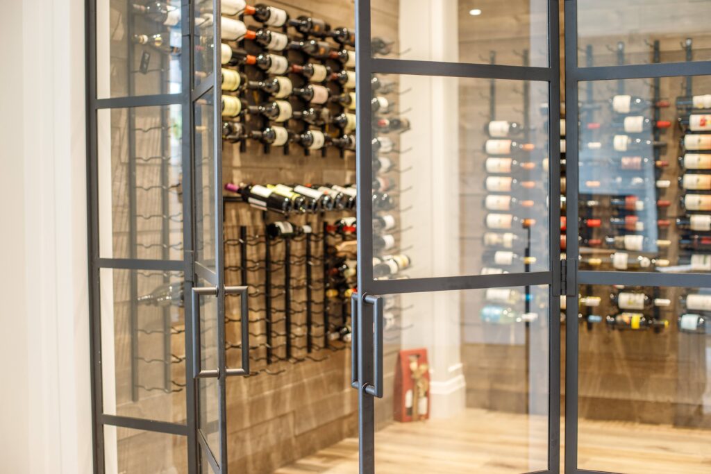A modern glass-enclosed wine cellar with metal racks holding various bottles of wine against wood-paneled walls on light hardwood flooring.